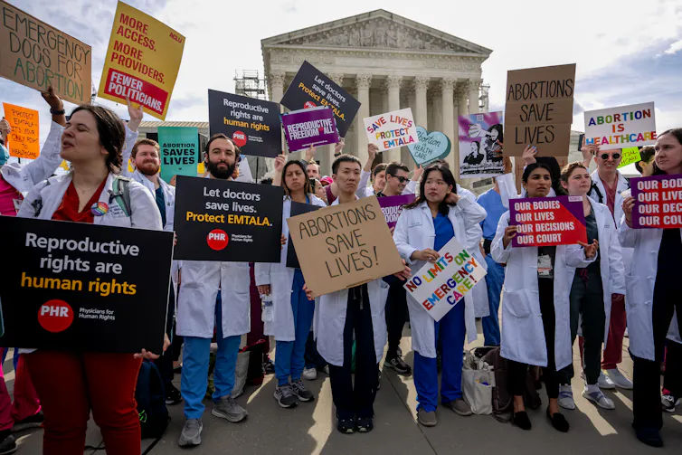 A group of people wear white doctors jackets and carry signs that say 'Emergency docs for abortion' and 'Reproductive rights are human rights.'