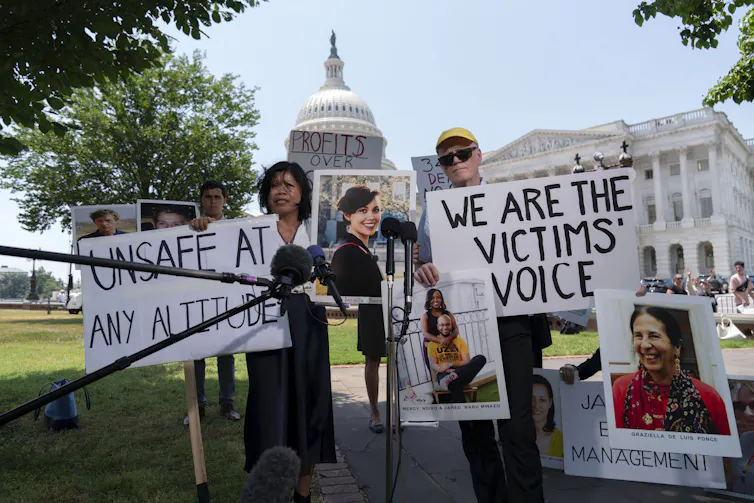 A group of people hold signs that say 'We are the victims' voice' and 'Unsafe at any altitude' while standing in front of the United States Capitol, a white neoclassical-style building