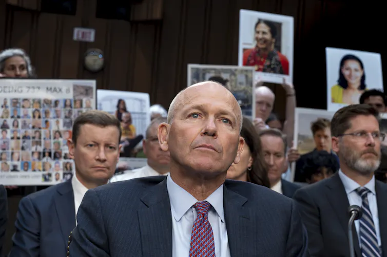A middle-aged bald white man in a suit sitting in front of a crowd of people holding protest signs
