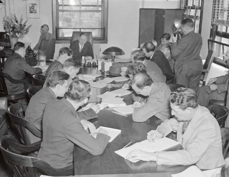 Une photo en noir et blanc montre plus d'une douzaine d'hommes rassemblés autour d'une longue table recouverte de documents.