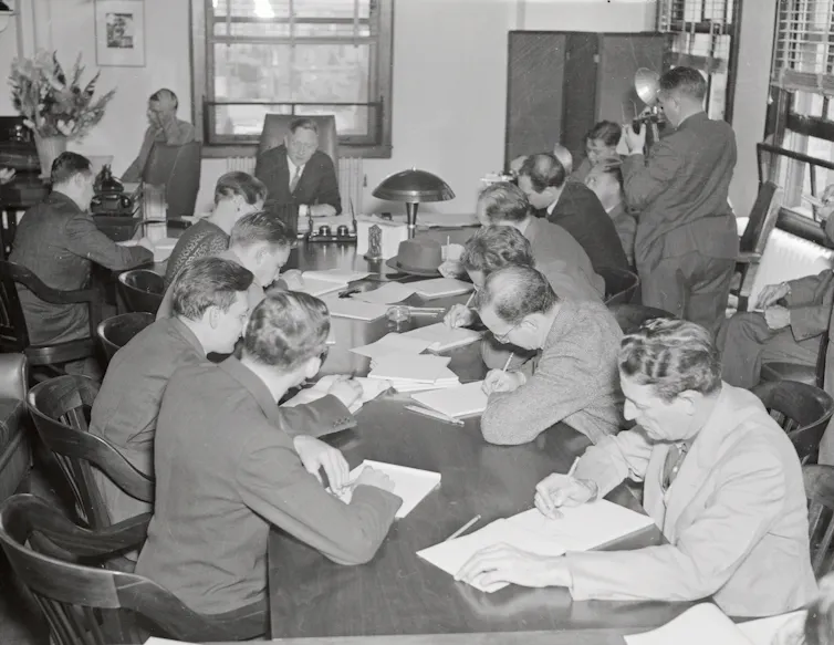 A black and white photo shows more than a dozen men gathered around a long table covered in documents.