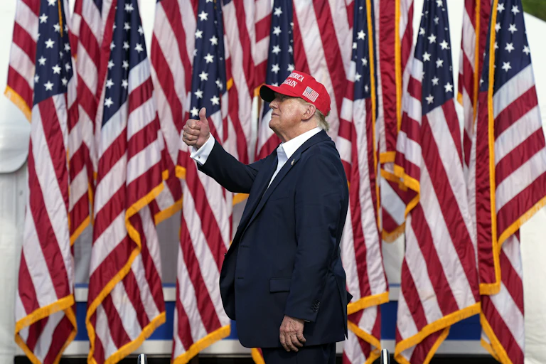 A man in a suit and a red hat signals 'thumbs-up' in front of a row of American flags.
