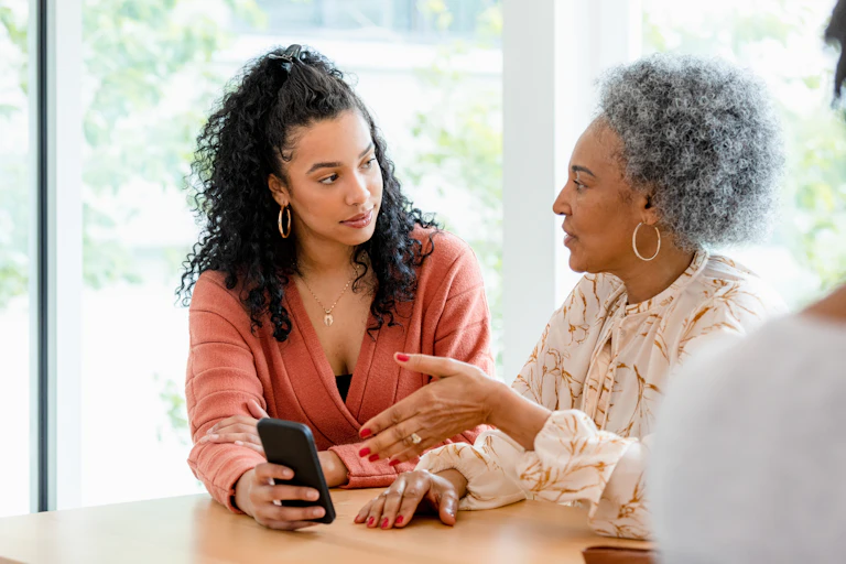 two seated women talking, one holds a phone and the other gestures