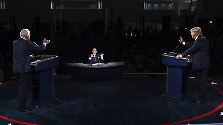Two men in dark suits at lecterns, speaking into microphone and gesturing.