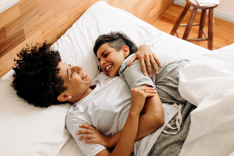 A queer couple cuddles in bed while laughing.