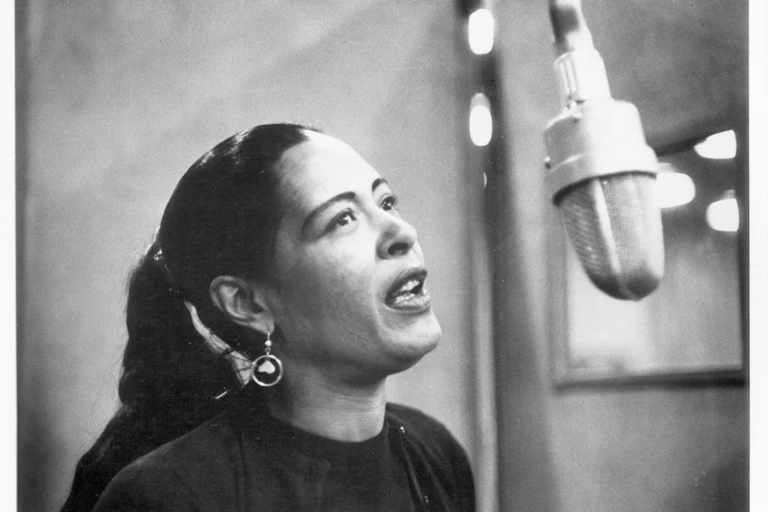 A black and white photo of a woman with hoop earrings and a long ponytail singing into a suspended microphone.