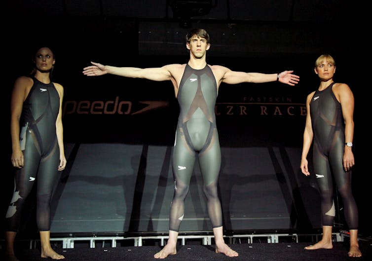 Three muscular athletes stand on a dark stage wearing one-piece charcoal coloured swimsuits.