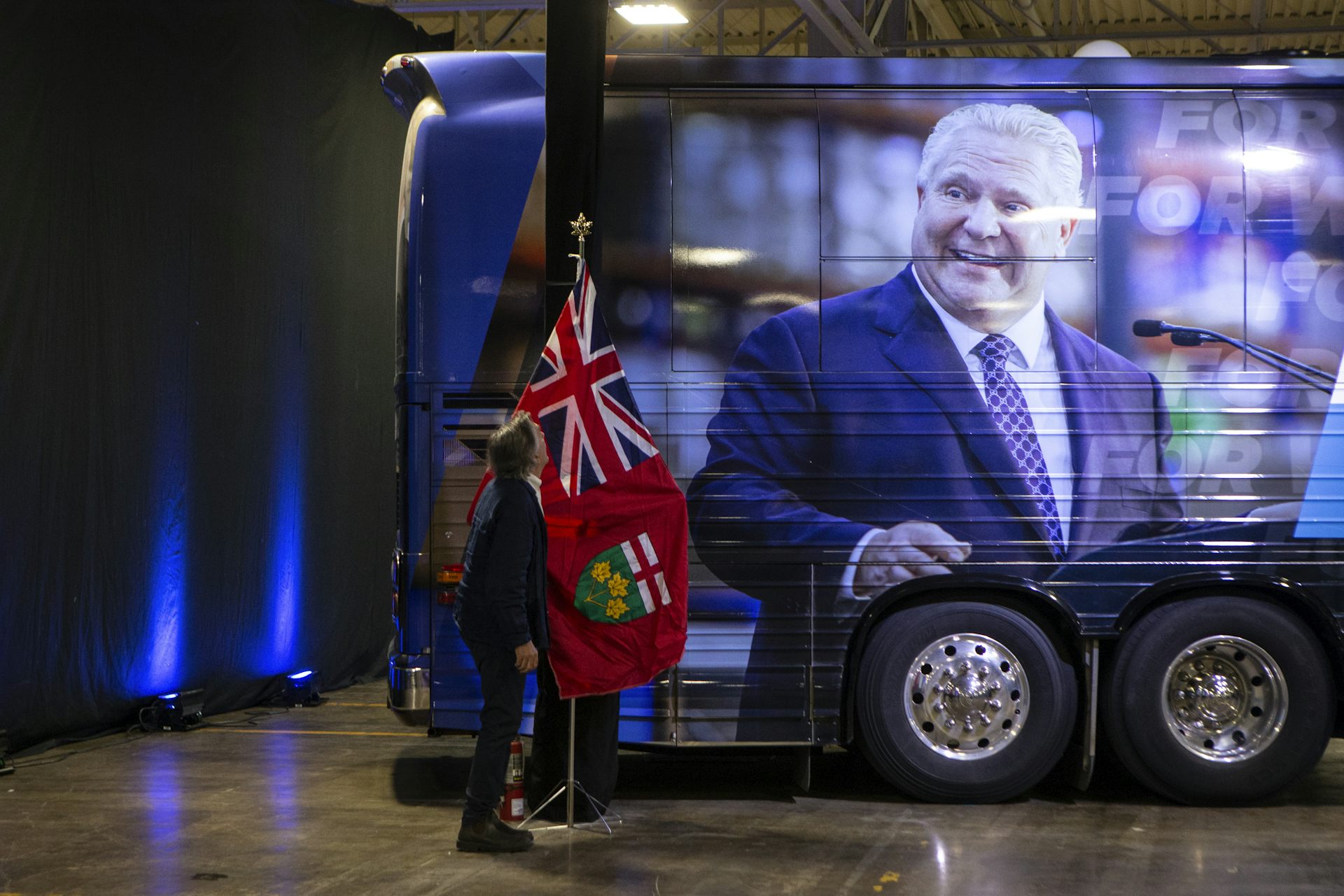 A man sets an Ontario flag next to a tour bus emblazoned with a photo of a smiling rotund man.