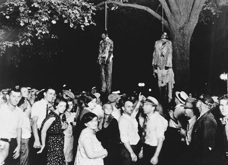 A black and white photograph of a crowd of people under a tree where two bloodied men have been hanged.