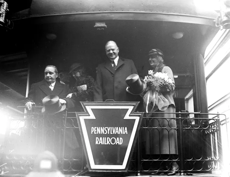 A man and a woman stand on a train balcony over a sign reading 'Pennsylvania Railroad.'