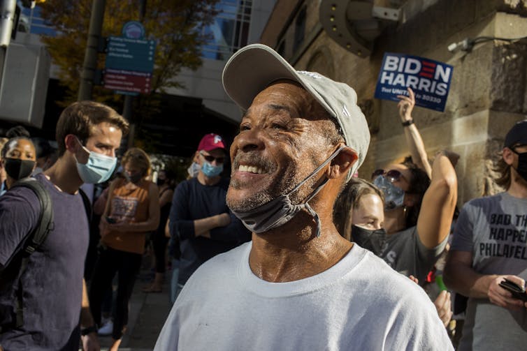 A man smiles widely in front of a crowd of celebrating people.