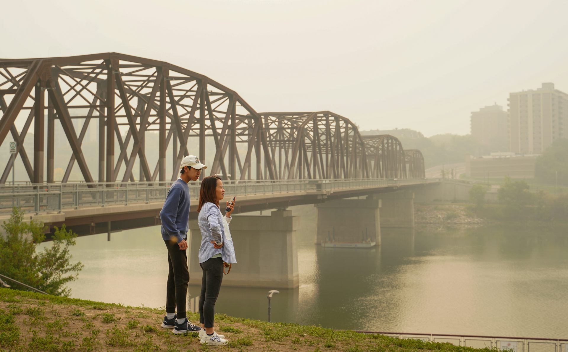 Two people by a bridge and river against a hazy, smoke-filled sky.