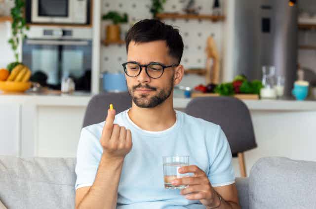 A man wearing glasses sitting on a sofa with a glass of water in one hand, looking at a pill in his other hand