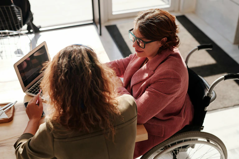 A woman in a wheelchair on a laptop talking to another woman.