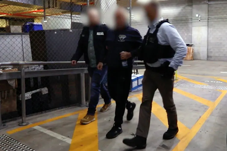 A man with his face blurred is escorted arm in arm by police officers, also with their heads blurred