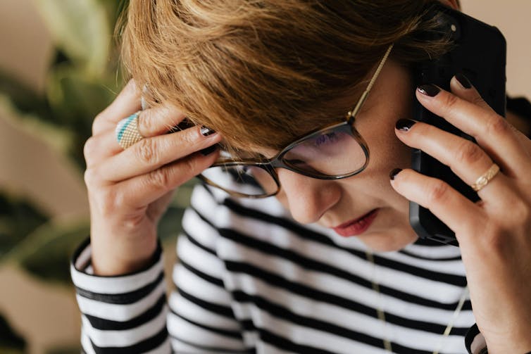 A woman holds a mobile phone up to her ear. She wears a serious expression and has her hand on her temple.
