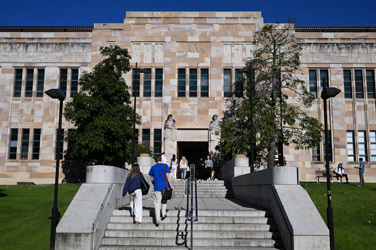 Young adults walk up stone steps to a main building, framed by trees.