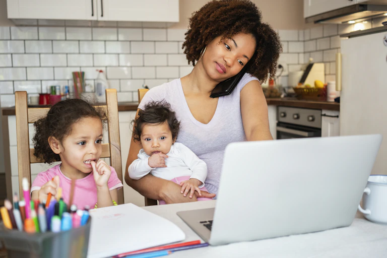 A woman works on her laptop while also talking on her phone, cradling a baby while a young child looks on. There are notes on the table