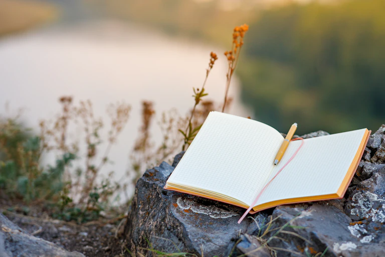 open empty book with pencil balanced on rock, blurry outdoor landscape in background