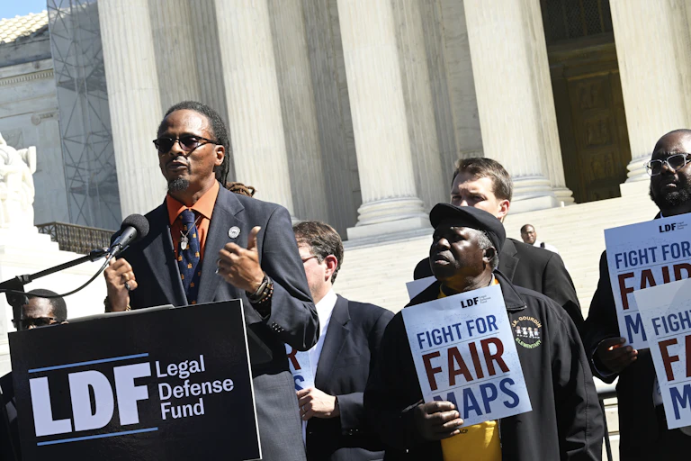 Black man at a podium with Supreme Court columns visible in background. Crowd holds signs reading 'fight for fair maps.'