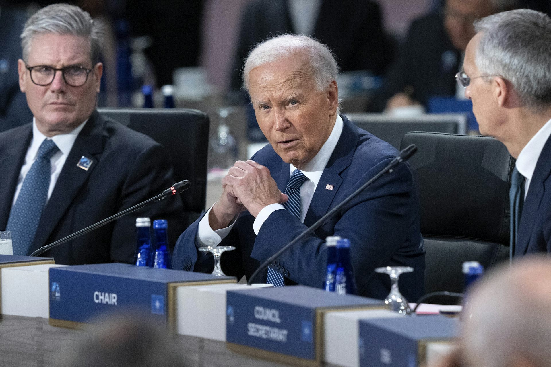 Three men with grey hair in dark suits chat behind a row of microphones.