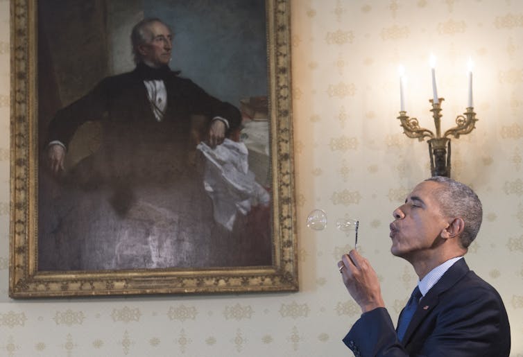 A man wearing a suit blows a bubble as he stands in front of an old looking oil portrait of a man.