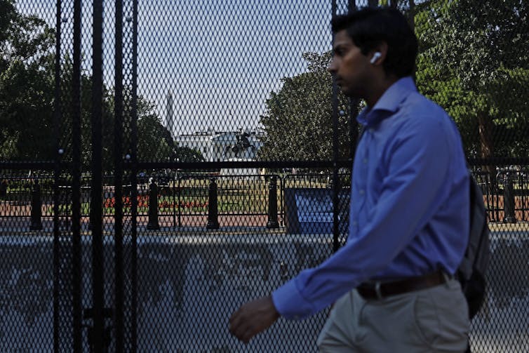 A man walks past a large black security fence. Behind the fence are a white building and trees.
