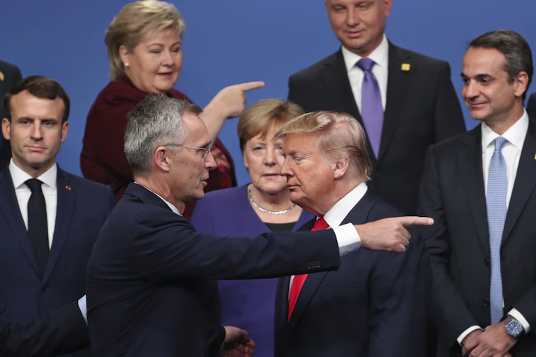 A man with grey hair and a blonde woman behind him point out of frame to a man who stands facing them with his eyes closed.