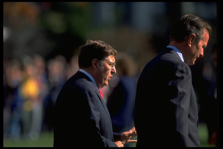 Two men wearing dark suits walk together, as seen from the side.