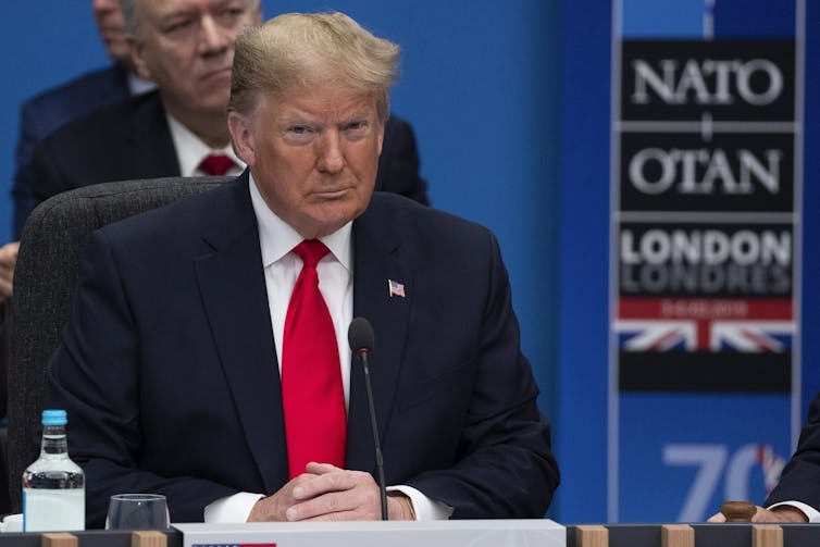A man with blond/white hair glowers while sitting at a meeting with a NATO sign behind him.