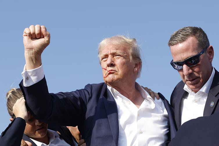 A white man with a bloodied face raises his fist as he is surrounded by men in darks suits and sun glasses.