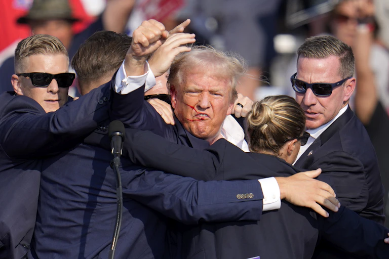 a man in a suit with blood streaks across his face raises a fist to the camera as three other men in navy suits and sunglasses surround him