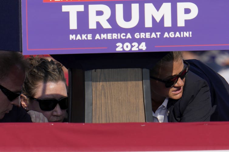 deux hommes en lunettes de soleil accroupis regardent autour d’eux, un panneau Trump violet est visible au premier plan