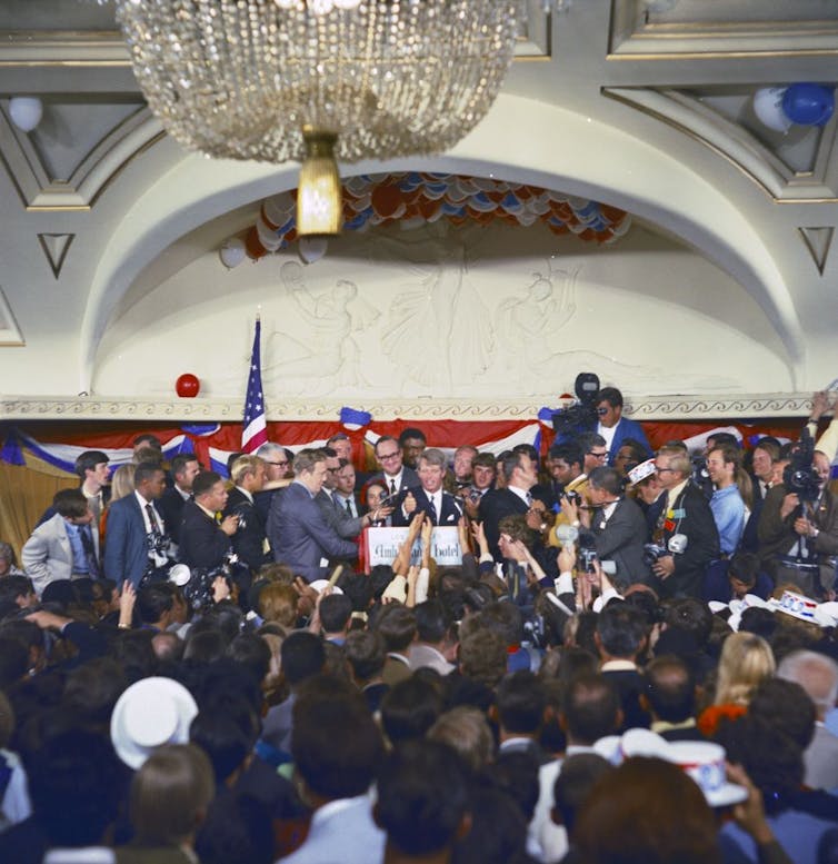 a man in a suit addresses a crowd in a ballroom