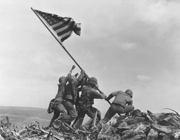 Black and white photograph, soldiers lean on each other to raise the US flag.