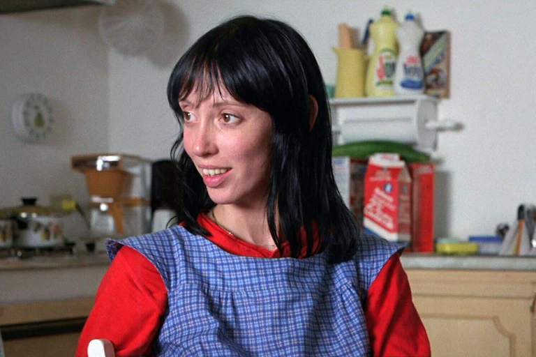 A young woman in red top and blue smock sitting in a a kitchen smiling andlooking off into the distance.