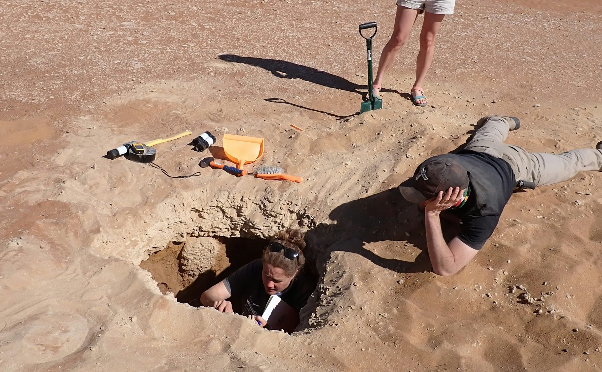The scientist sits in a small hole studying the sediment and taking samples while his colleagues look on.
