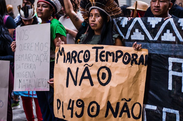 Manifestantes vestindo cocares indígenas seguram cartazes contra a teoria jurídica do Marco Temporal