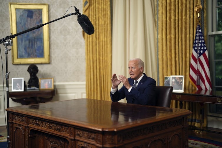 An aging man sits at an ornate wooden desk with gold curtains behind him. A boom microphone is over his head.