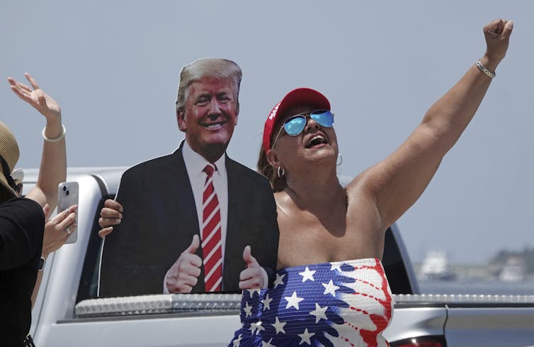 A woman in a stars-and-stripes tube top and red ball cap stands beside an cardboard cutout of Trump and cheers.