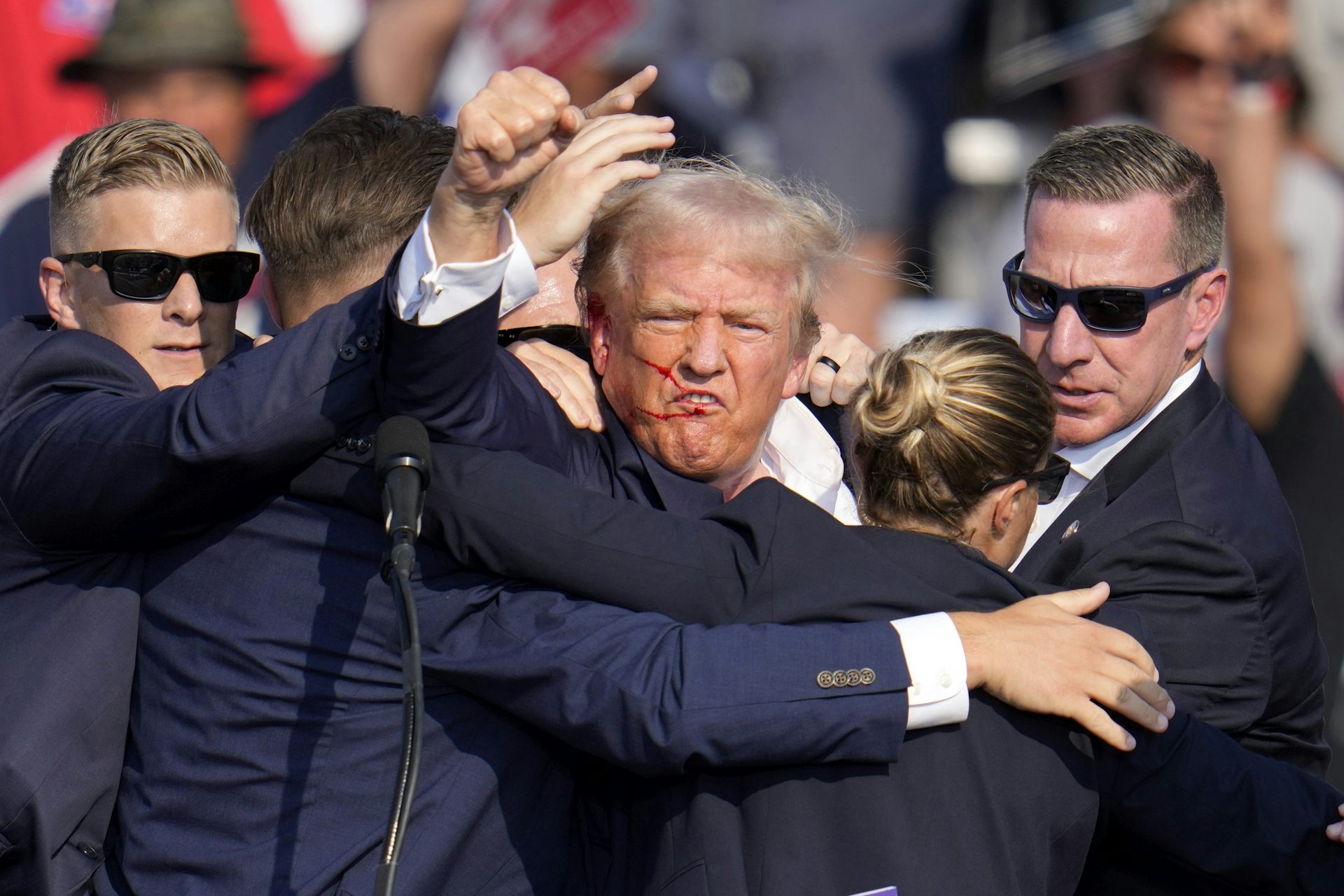A man with greyish blond hair and a streak of blood on his face raises his fist in the middle of a huddle of Secret Service agents.