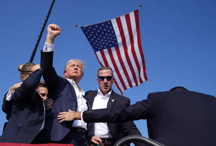 A man with greyish blond hair and a streak of blood on his face raises his fist in the middle of a huddle of Secret Service agents. An American flag flies next to him.