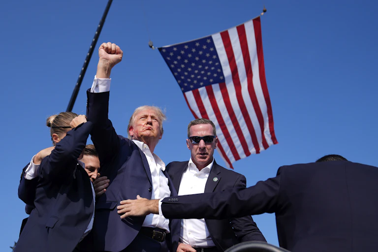 A man with greyish blond hair and a streak of blood on his face raises his fist in the middle of a huddle of Secret Service agents. An American flag flies next to him.