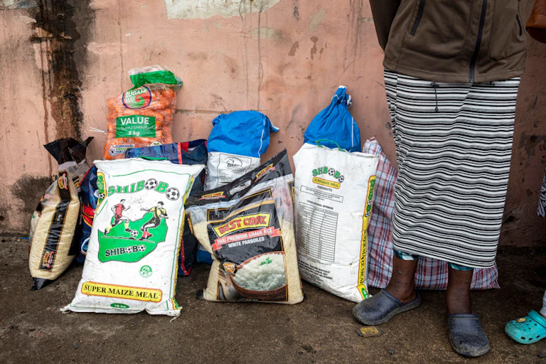 Large bags of maize meal, rice and carrots propped against a wall, with a woman standing next to them
