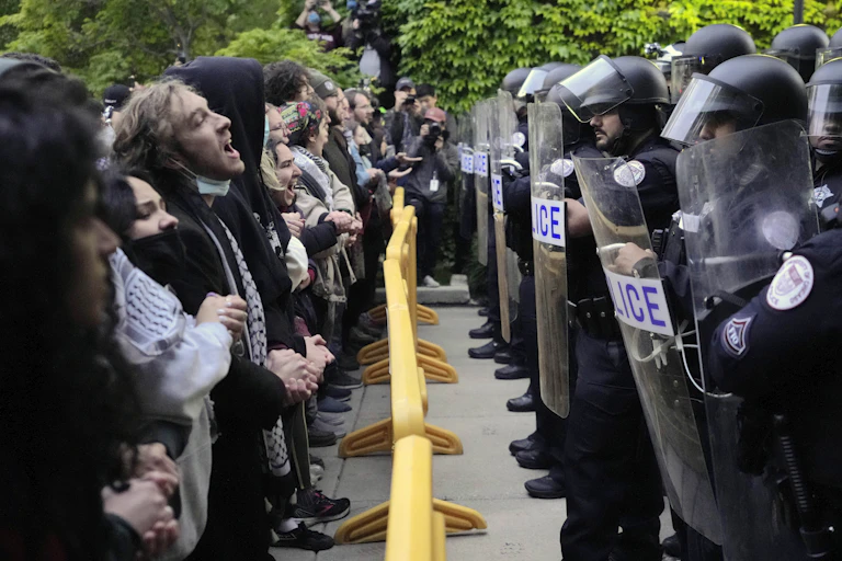 line of young people on the left, some wearing keffiyah scarves, face a row of riot police holding shields. They are separated by a yellow metal barrier.