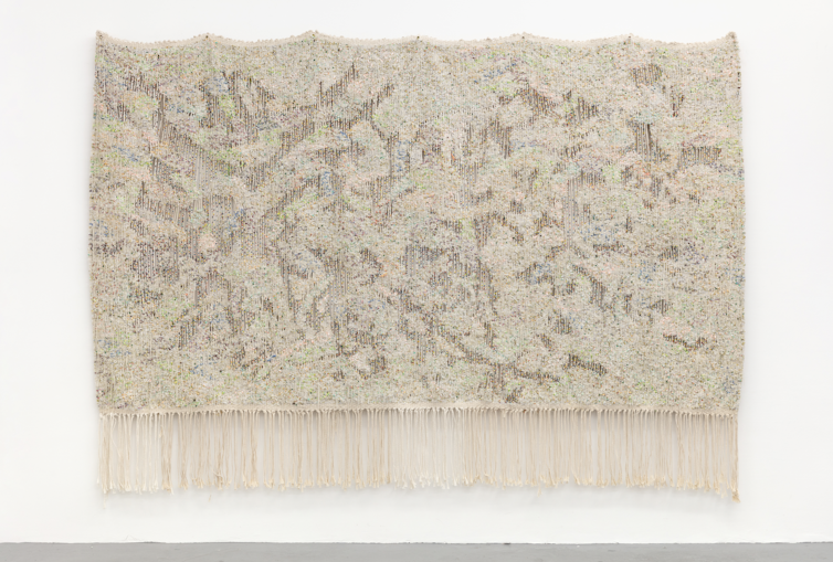 A white tapestry covered in marks hanging on a gallery wall.