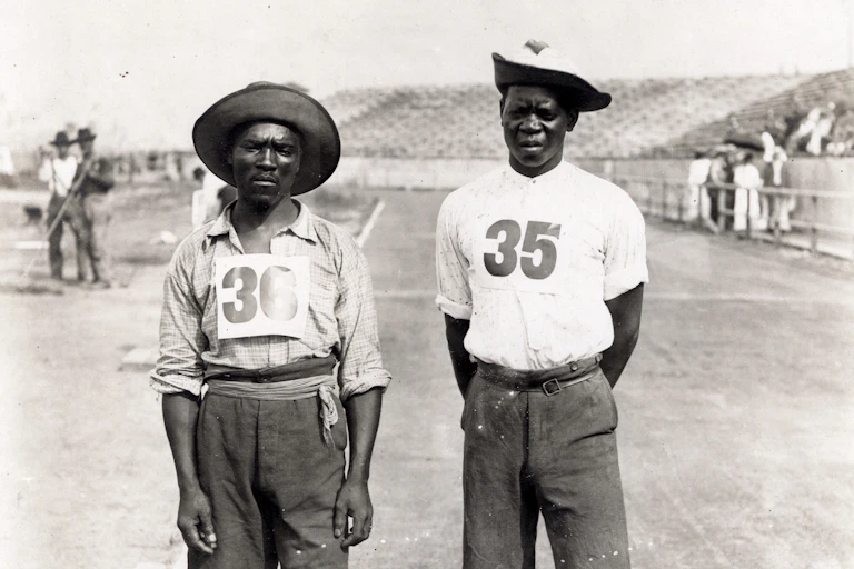 A vintage black and white photo of two men standing on a running track, wearing the numbers 35 and 36 on their shirts, ragged shorts and floppy hats.