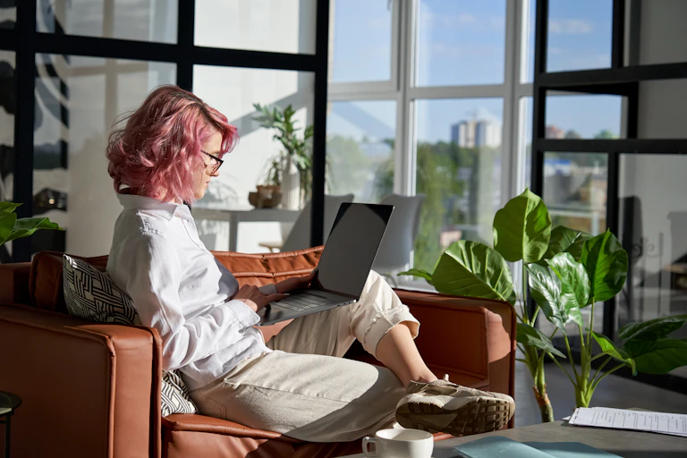 A young woman with pink hair and a stylish outfit sits on a sofa in a cool, bright office space, working on a laptop