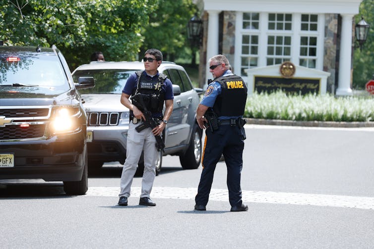 Two people wearing black vests and holding guns stand near two parked SUVs on an otherwise empty street in the daytime.