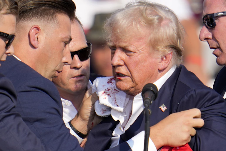 An older white man with white hair stands at a microphone, surrounded by men in dark blue suits. One of them holds a white towel up to his face and there is blood on it.